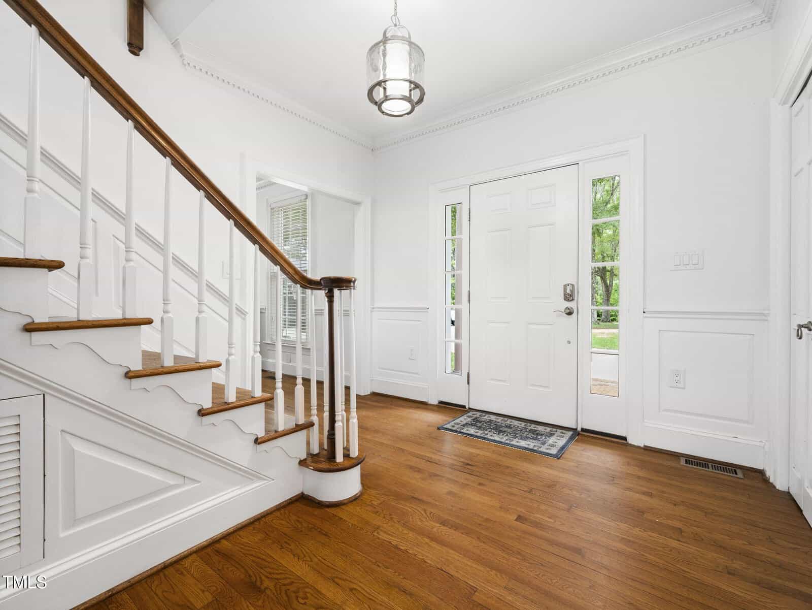 A bright entryway with wooden floors, a white front door with sidelights, a staircase with white spindles and wooden handrail, white walls with decorative molding, and a modern hanging light fixture.