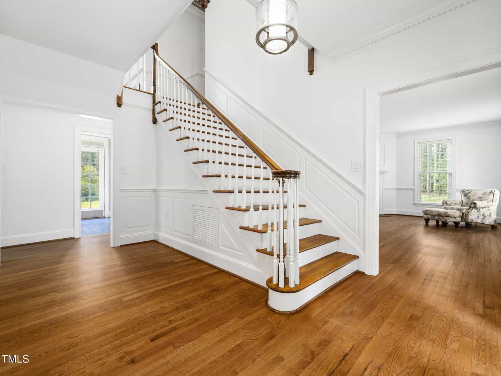 Bright entryway with wooden stairs, white banister, hardwood floors, and white walls. A large light fixture hangs from the ceiling, and two open doorways lead to sunlit rooms with large windows and classic trim.