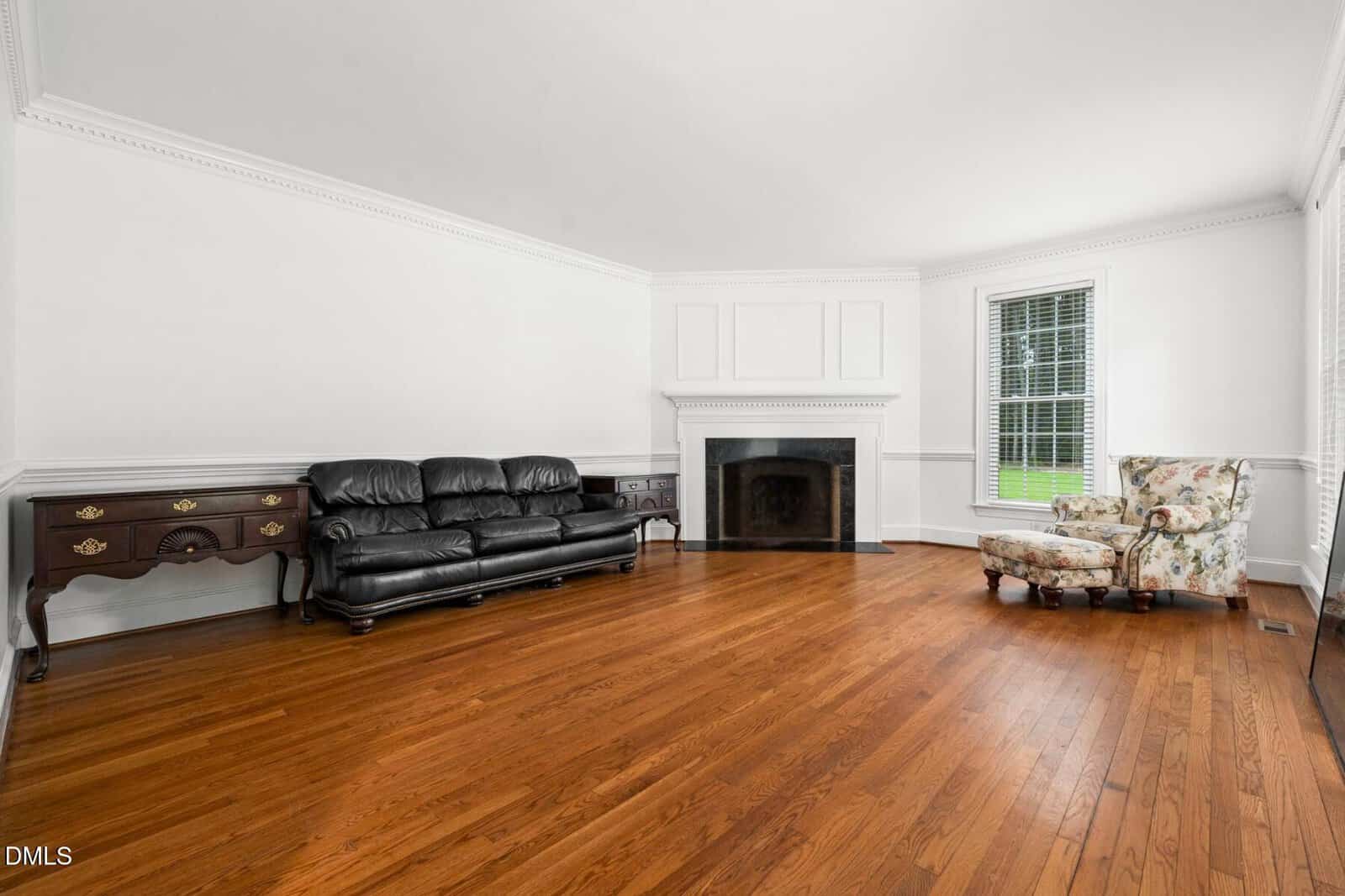 A spacious living room with hardwood floors, a black leather sofa, two wooden side tables, a fireplace with a white mantel, a floral armchair, and a large window letting in natural light.