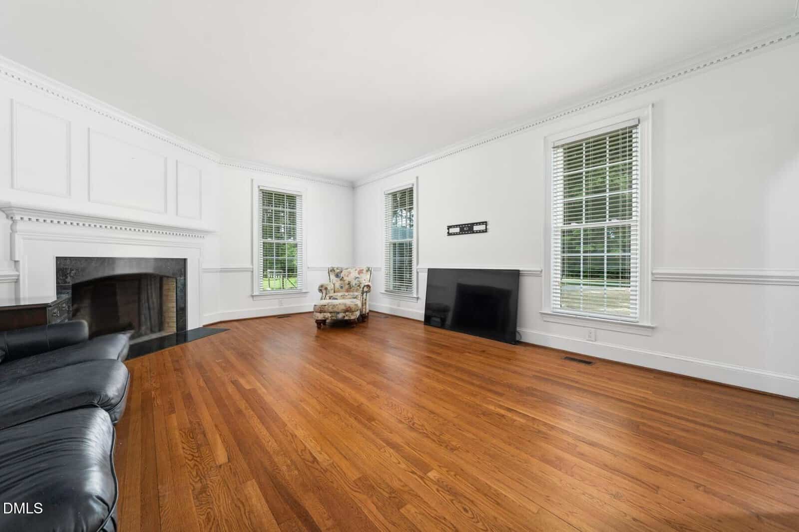 A spacious, bright living room with white walls, two tall windows, a black fireplace, hardwood floors, a floral armchair, a black sofa, and a large flat-screen TV leaning against the wall.