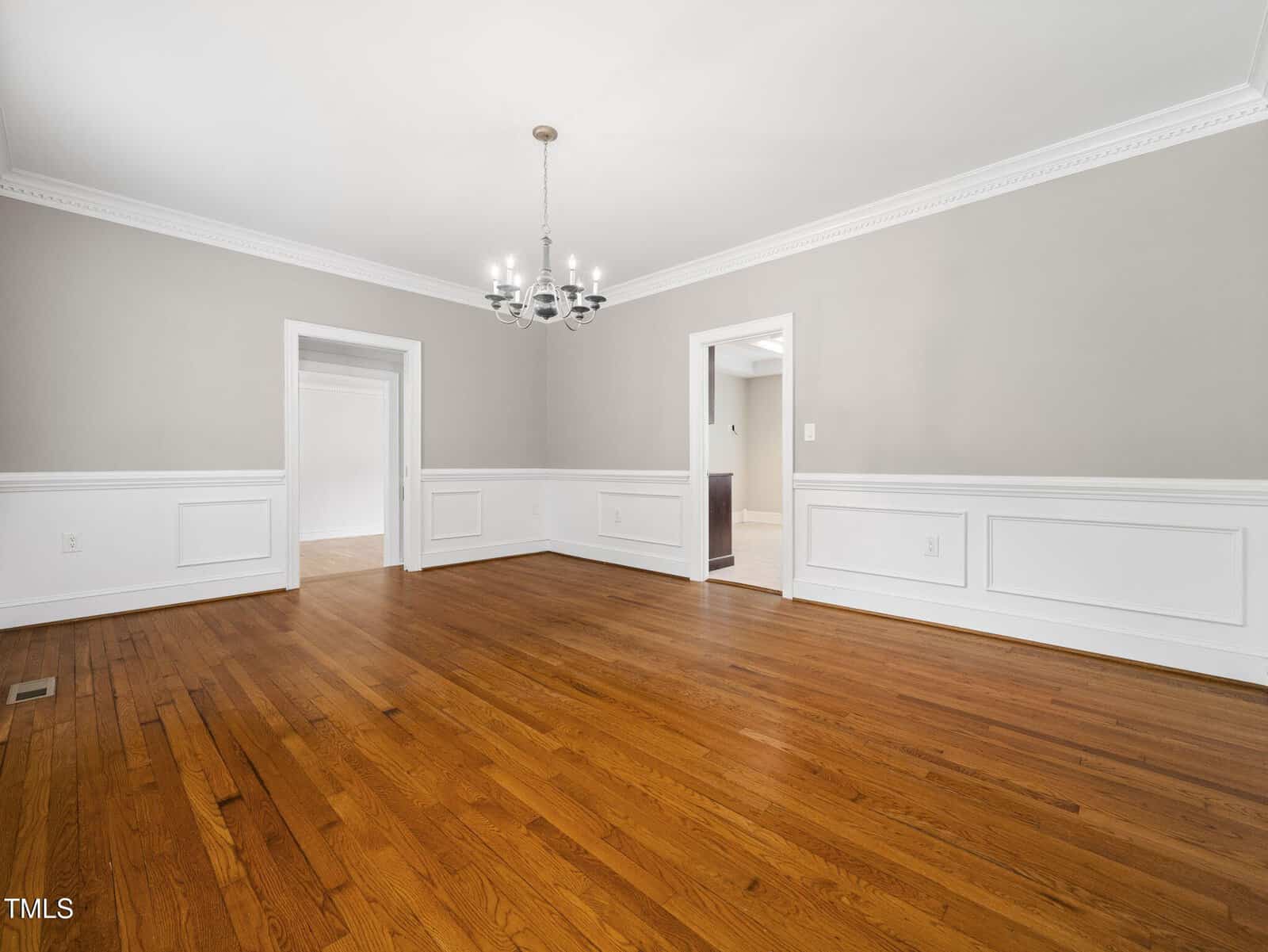 A spacious, empty dining room with hardwood floors, light gray walls, white wainscoting, a white ceiling, and a modern chandelier. Two doorways lead to adjacent rooms.
