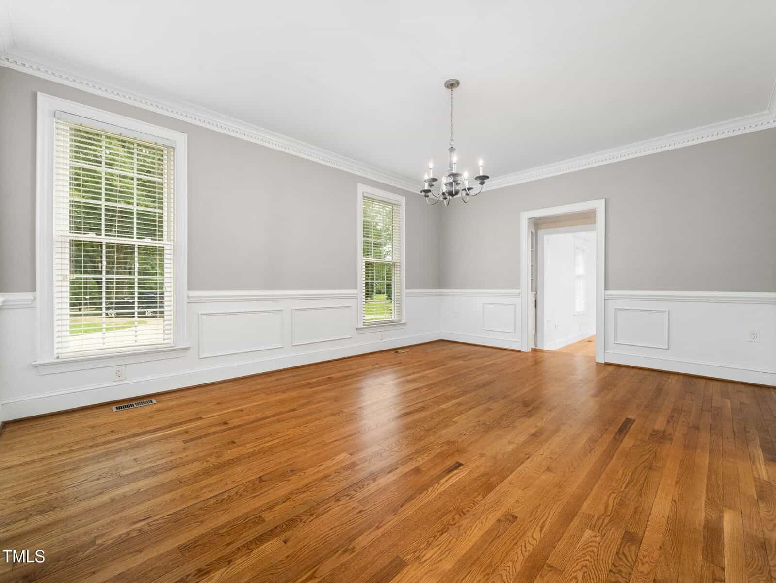 A spacious, empty room with light gray walls, white trim and wainscoting, large windows with blinds, a wooden floor, and a chandelier hanging from the ceiling. An open doorway leads to another bright room.