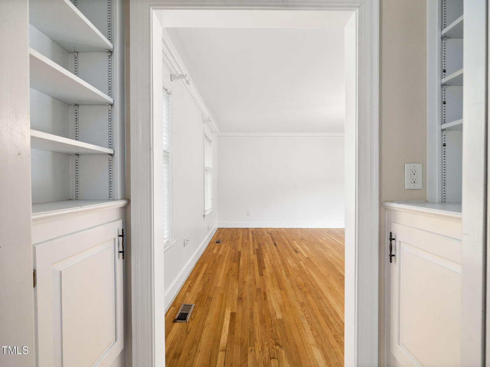 View through a doorway into an empty, bright room with polished hardwood floors, white walls, and trim. Built-in white shelves and cabinets are visible on both sides of the doorway.