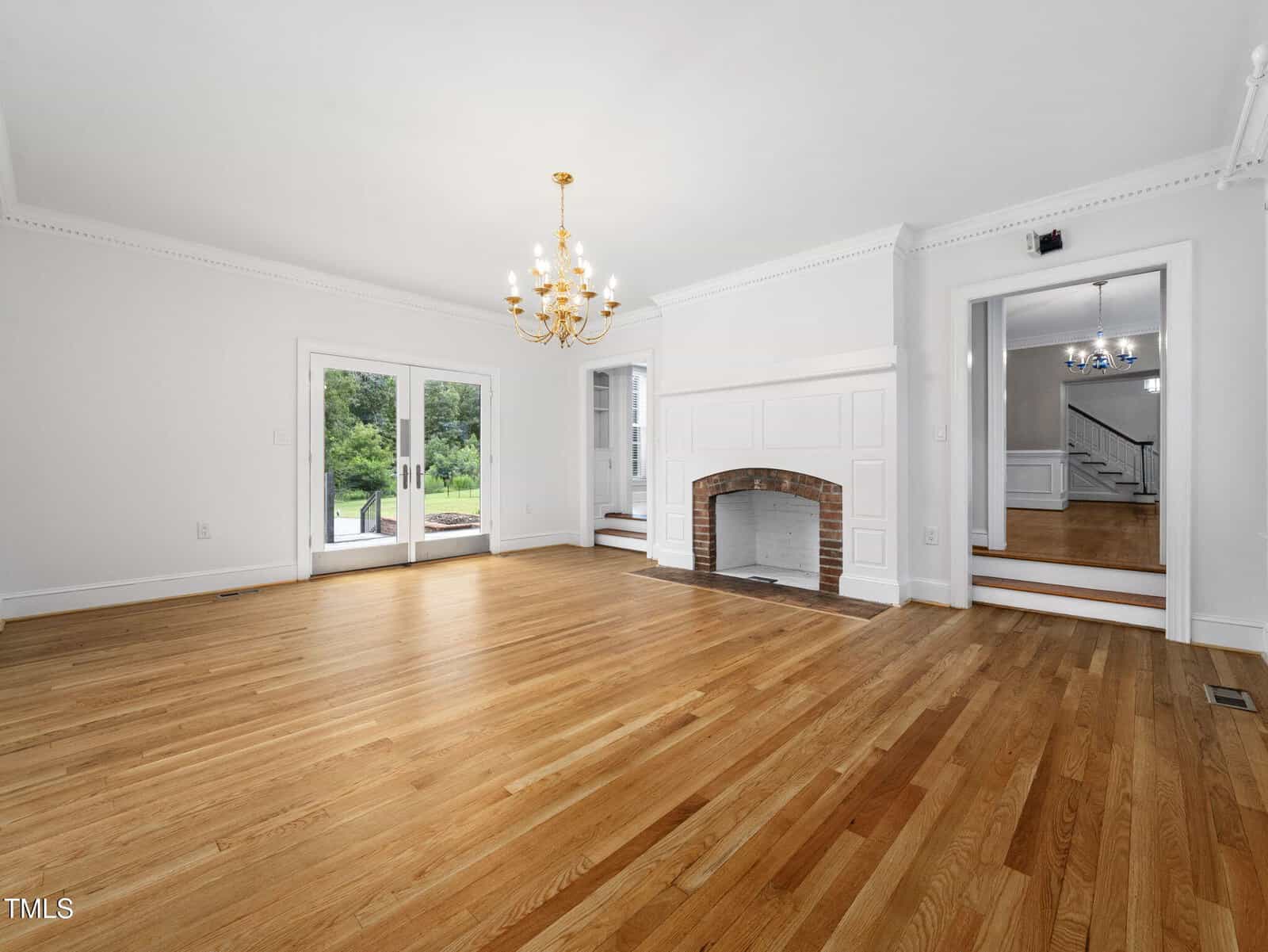 Spacious, empty living room with light hardwood floors, white walls, a decorative fireplace, gold chandelier, and large glass doors opening to a green outdoor area. Steps lead to an adjoining room.