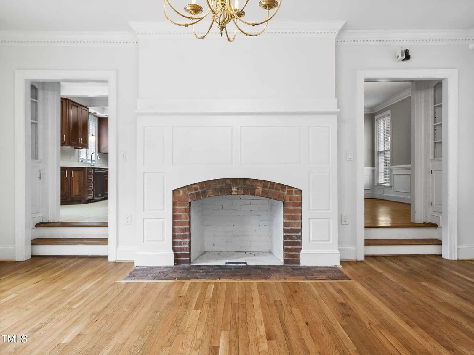 A bright room features a white fireplace with a brick interior, hardwood floors, a gold chandelier, and two doorways leading to a kitchen on the left and a dining area on the right.