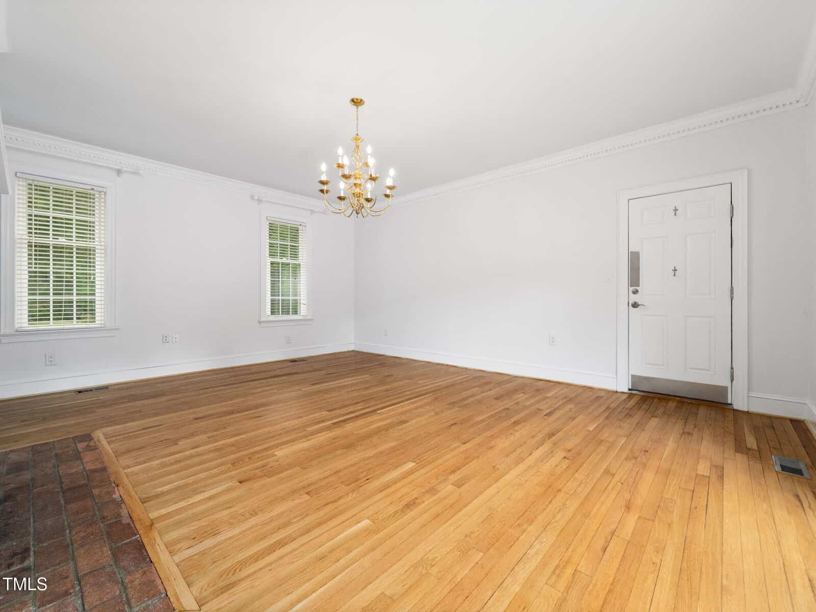 Empty living room with hardwood floors, white walls, two windows with blinds, a white front door, a gold chandelier, and a small brick section in the corner.