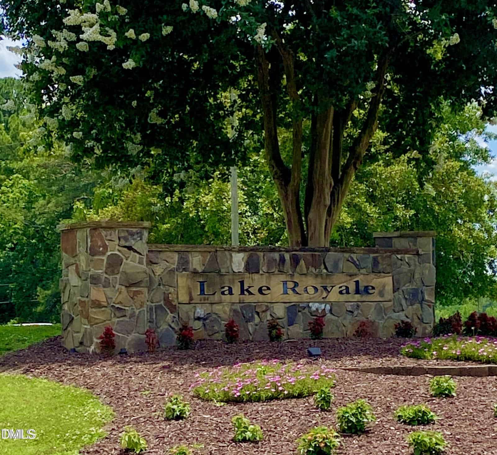 A stone entrance sign reads "Lake Royale" at 100-106 Cherokee Drive, Louisburg, surrounded by a mulched area with small plants and flowers. A large tree and green foliage create a scenic backdrop under a partly cloudy sky.