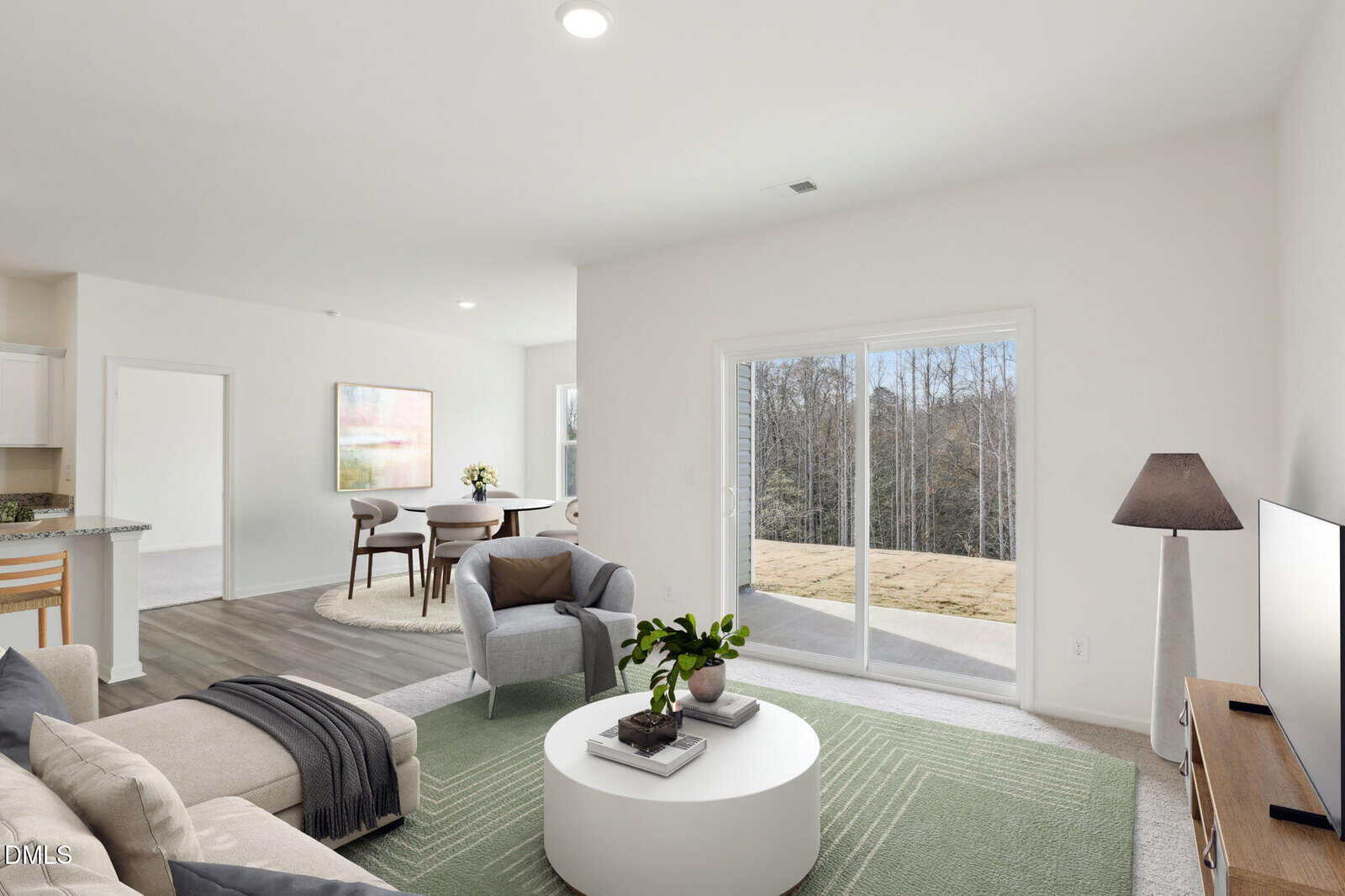 A bright, modern living room with a green area rug, beige sofa, armchair, round coffee table, standing lamp, TV, and sliding glass doors leading to a patio with a wooded view. Dining area and kitchen are visible in the background.
