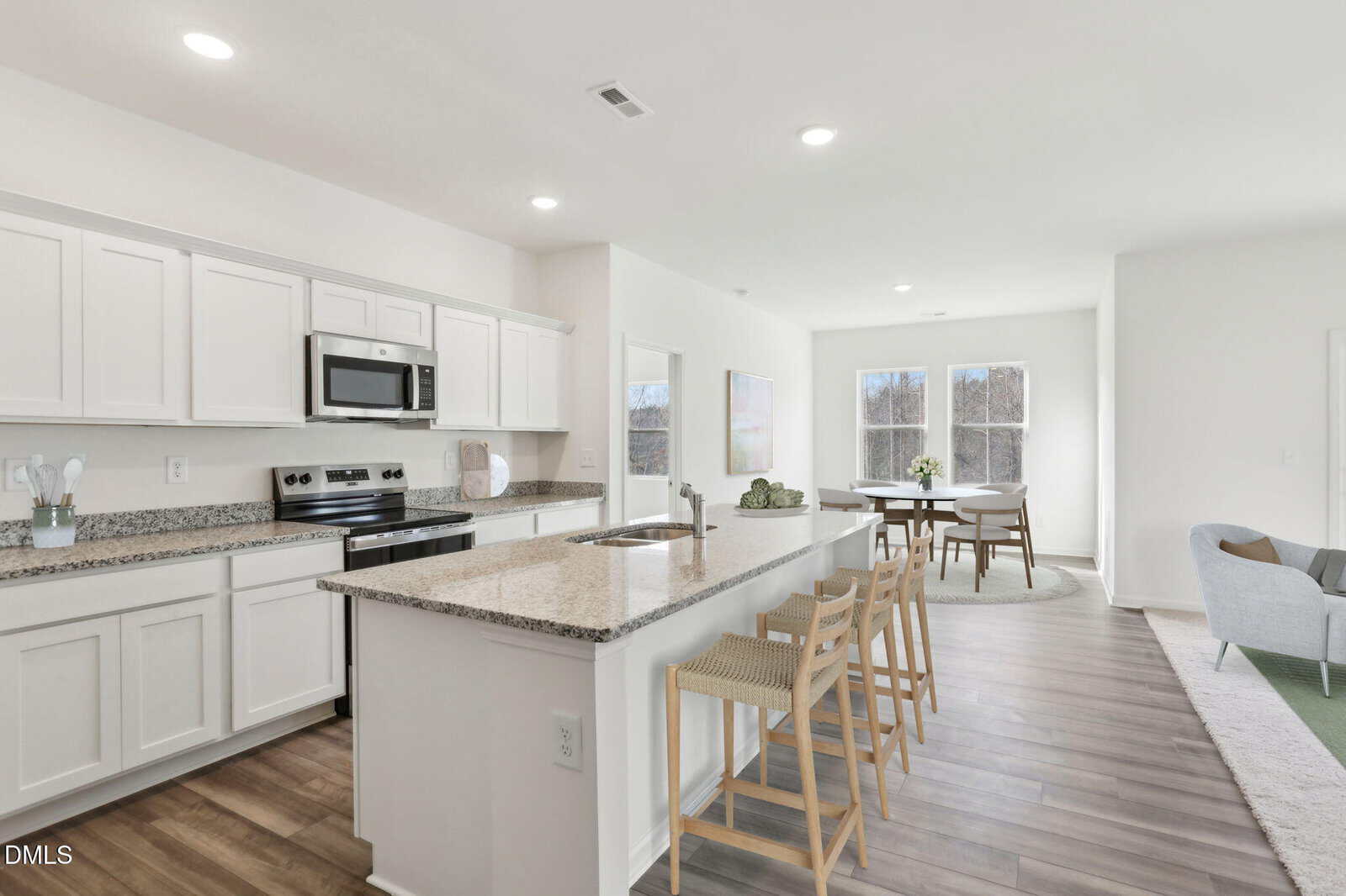 Modern kitchen with white cabinets, stainless steel appliances, a granite countertop island with three wooden bar stools, and an open layout leading to a dining area and living room with large windows.