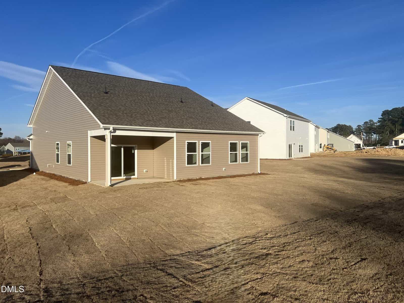 Newly constructed beige house with a covered patio, multiple windows, and a large dirt backyard. Other similar homes are visible nearby under a clear blue sky.