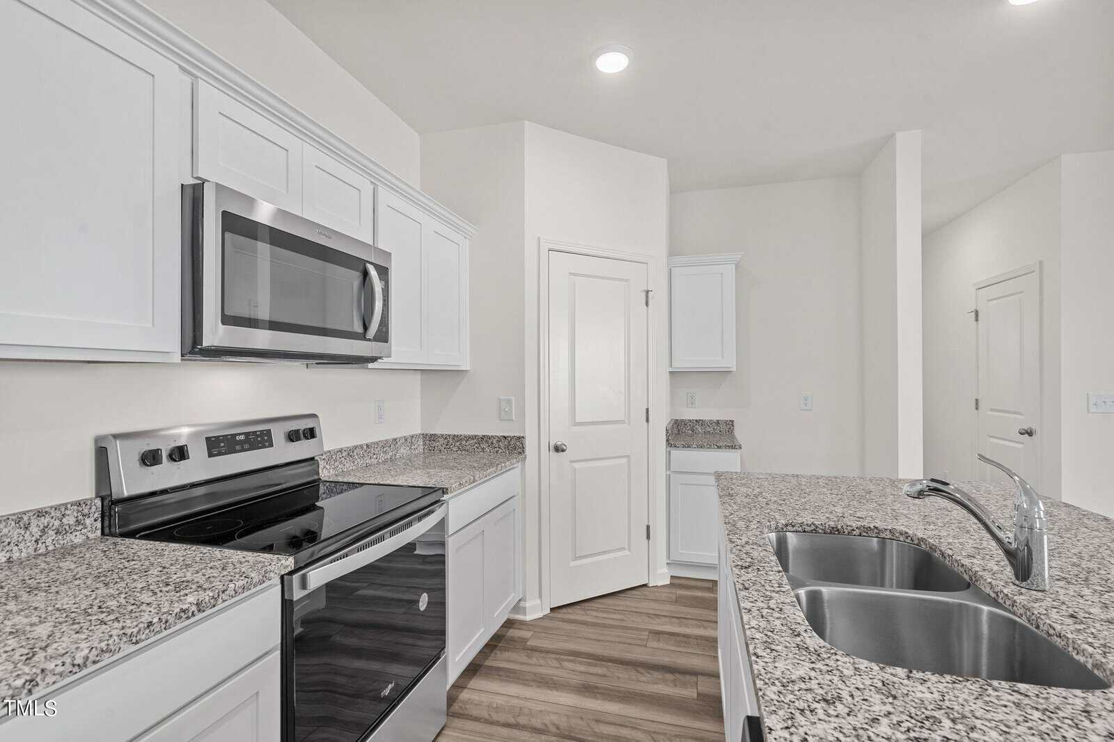 Modern kitchen with white cabinets, stainless steel appliances, granite countertops, double sink, and wood-style flooring. The room is well-lit and features a central pantry with a white door.