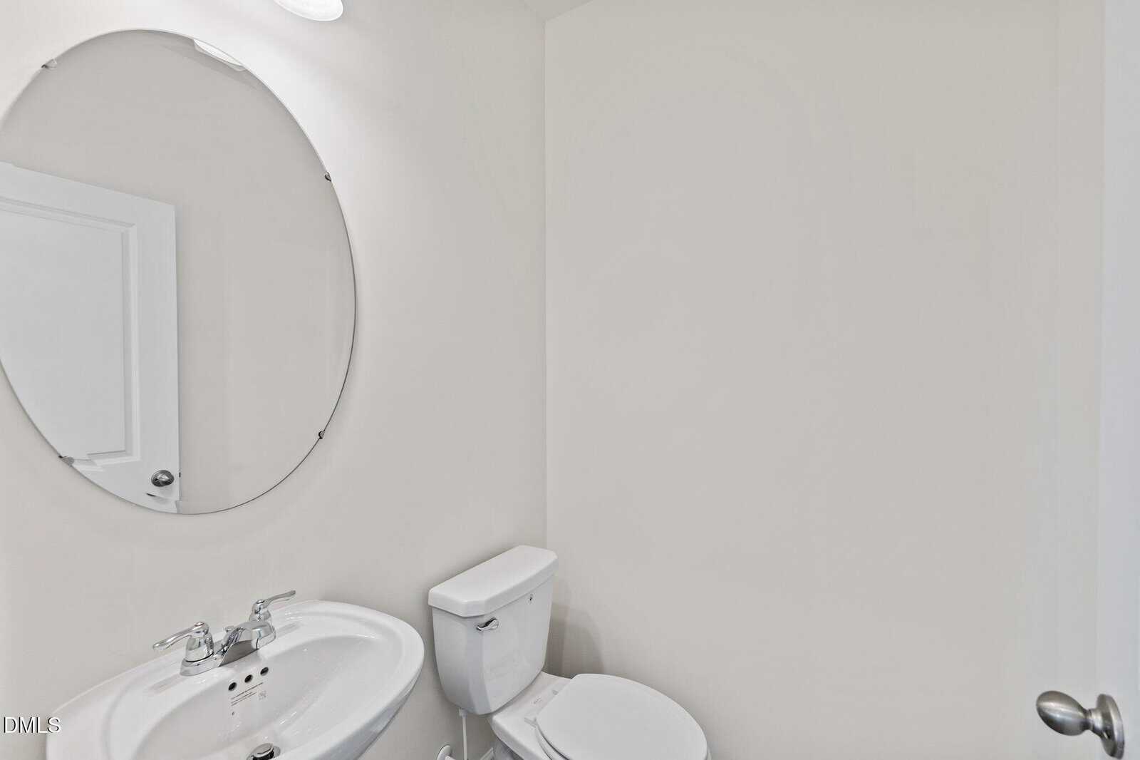 A small, minimal bathroom with white walls, a white pedestal sink with a chrome faucet, a round mirror above the sink, and a white toilet. The space is clean and simple with modern fixtures.