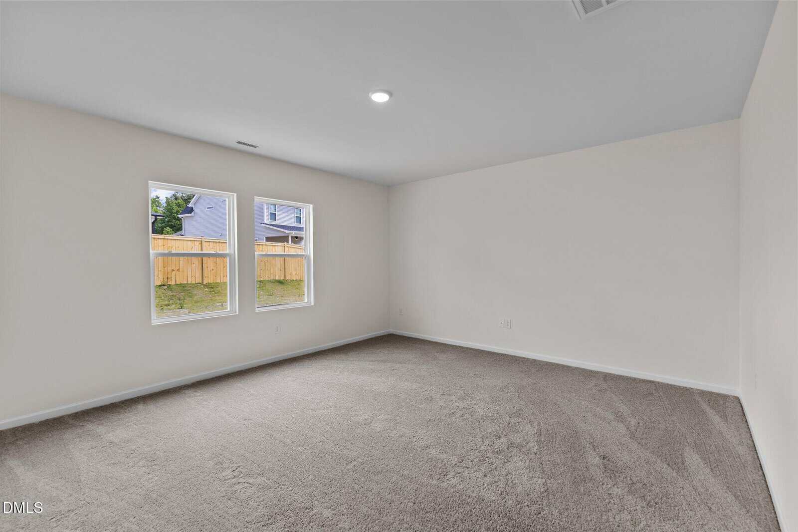 A spacious, empty room with beige walls, gray carpet, and two windows letting in natural light. A wooden fence and neighboring house are visible outside. The ceiling has a recessed light fixture.