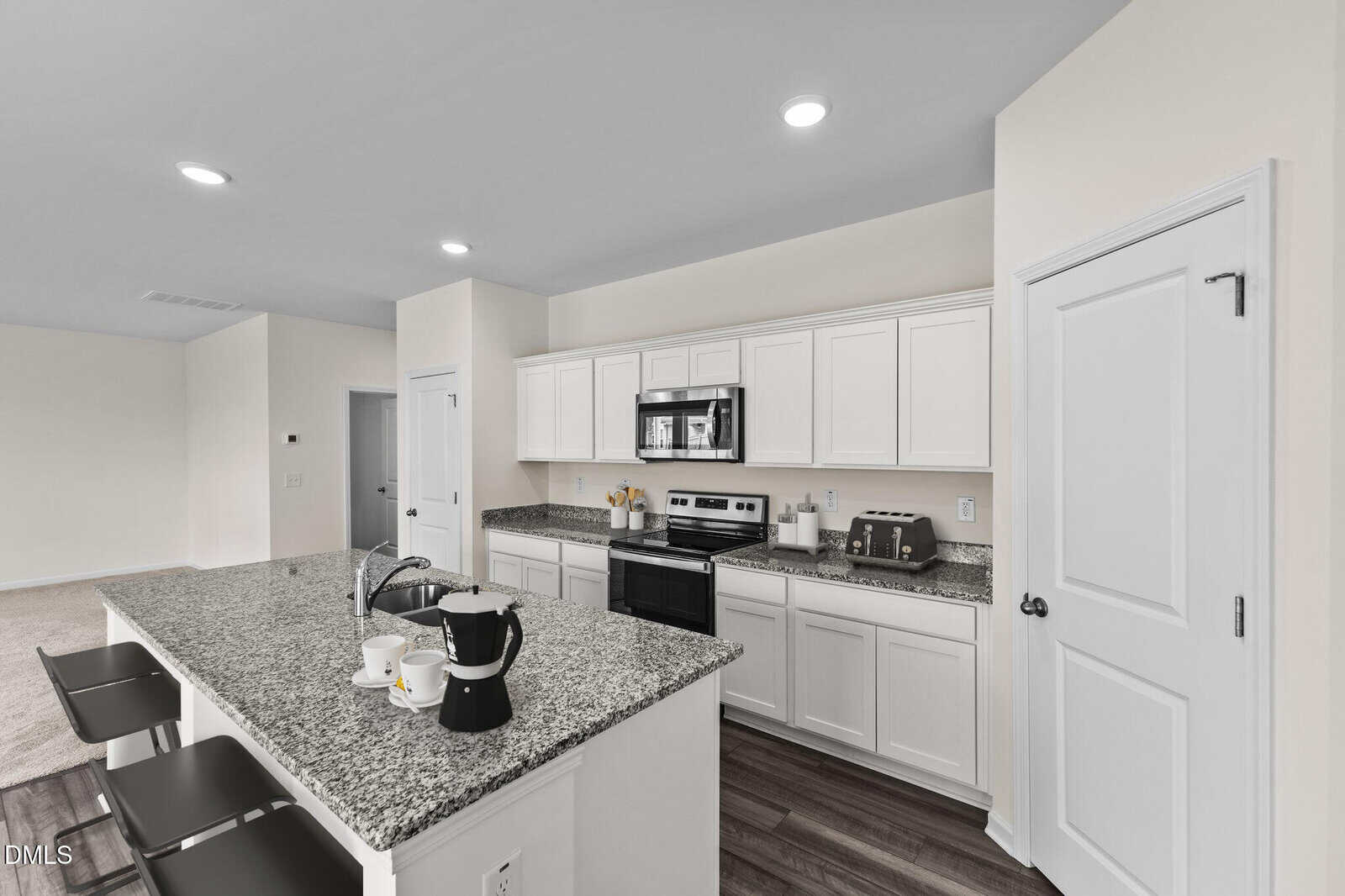 Modern kitchen with white cabinets, granite countertops, stainless steel appliances, and a large island with barstools. Coffee mugs and a coffee maker sit on the island. The space is bright and open.
