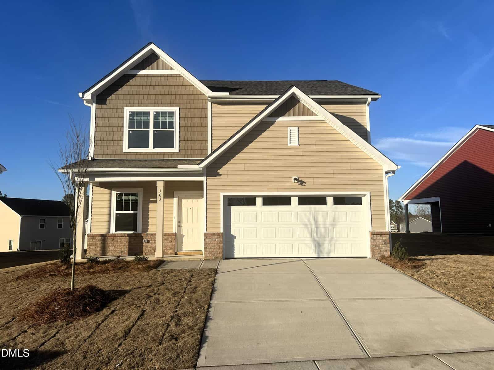Two-story tan house with a double garage, white trim, and a porch on the left, located at 103 Big Dream Court in Louisburg. The yard has new landscaping, and the sky is clear and blue.