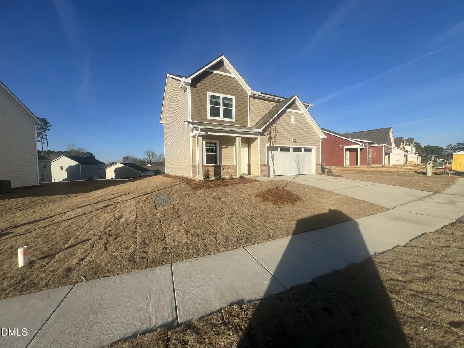 A two-story tan house with a front porch, attached garage, and a sloped yard with sparse grass. The home is part of a suburban neighborhood with clear skies and a concrete sidewalk in front.