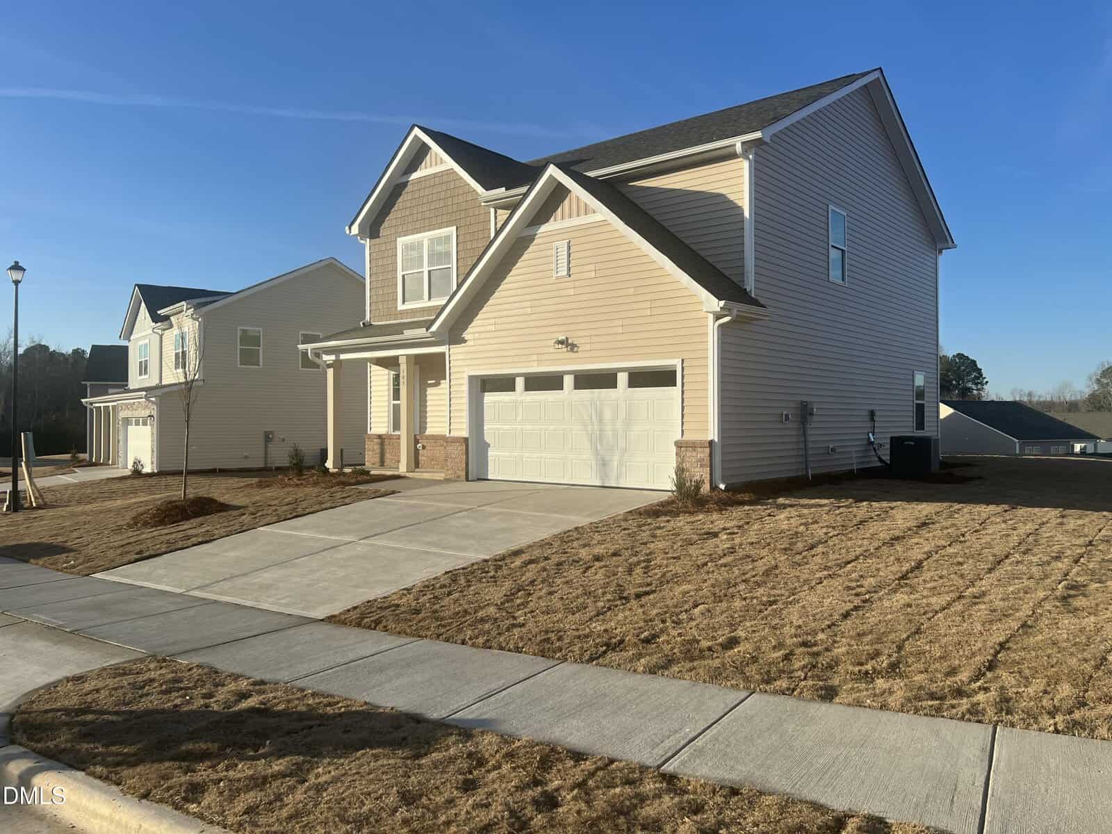A modern two-story beige house with a double garage sits on a corner lot with a freshly mowed lawn under a clear blue sky in a suburban neighborhood.