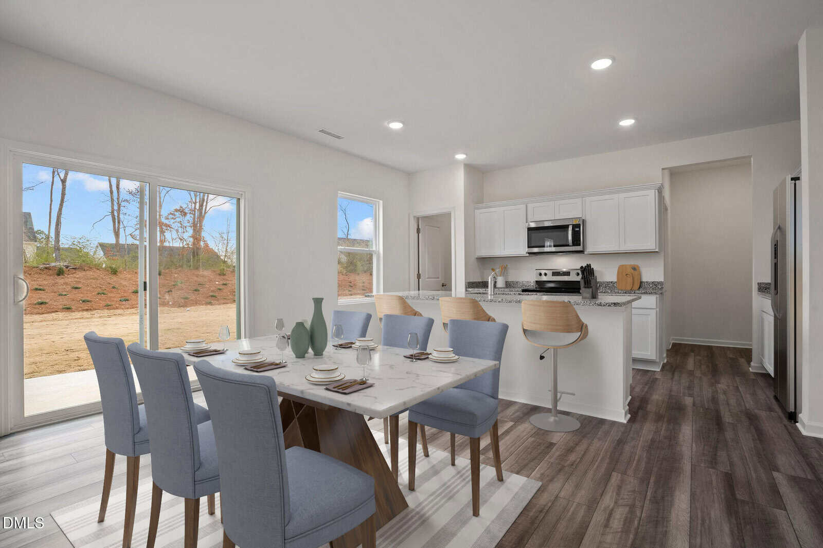 A modern dining area with a marble table and blue chairs sits next to a kitchen with white cabinets, stainless steel appliances, bar stools, and large windows overlooking a backyard.