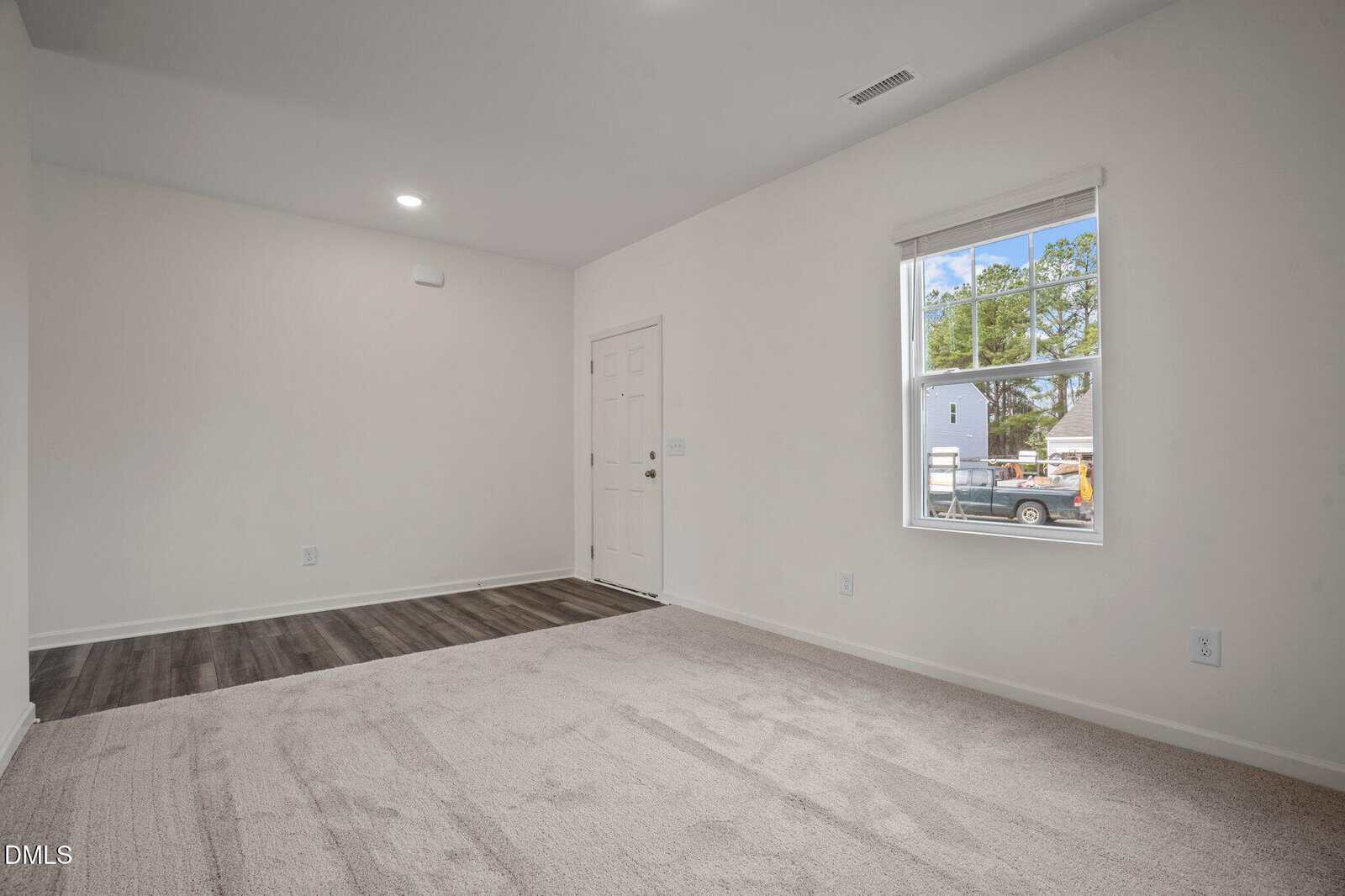 A bright, empty room with beige carpet, white walls, a window with a view outside, a white door, and a section of dark wood flooring near the entrance.