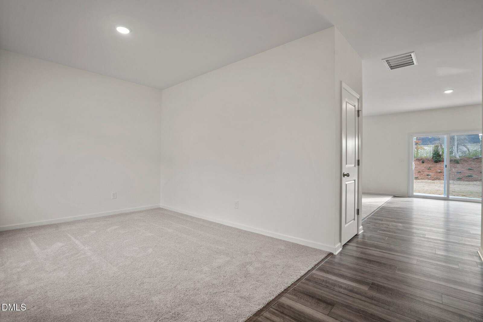A room with light beige carpet on the left, white walls, and a door leading to a wood-floored area. Sliding glass doors at the back open to a yard with mulch and grass. Ceiling lights illuminate the space.
