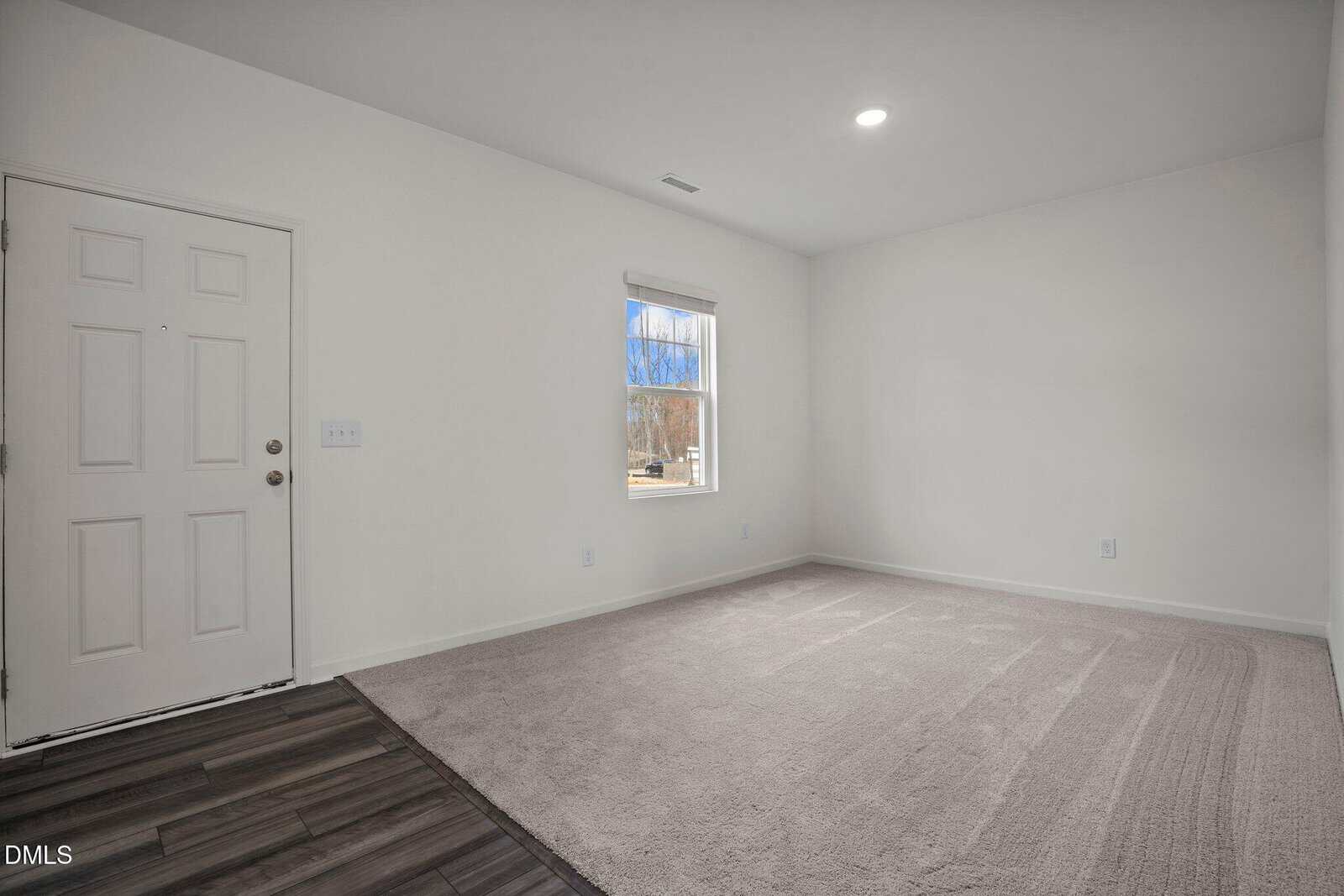 A bright, empty room with white walls, a gray carpet, a window letting in natural light, a closed white door, and dark wood flooring near the entrance.
