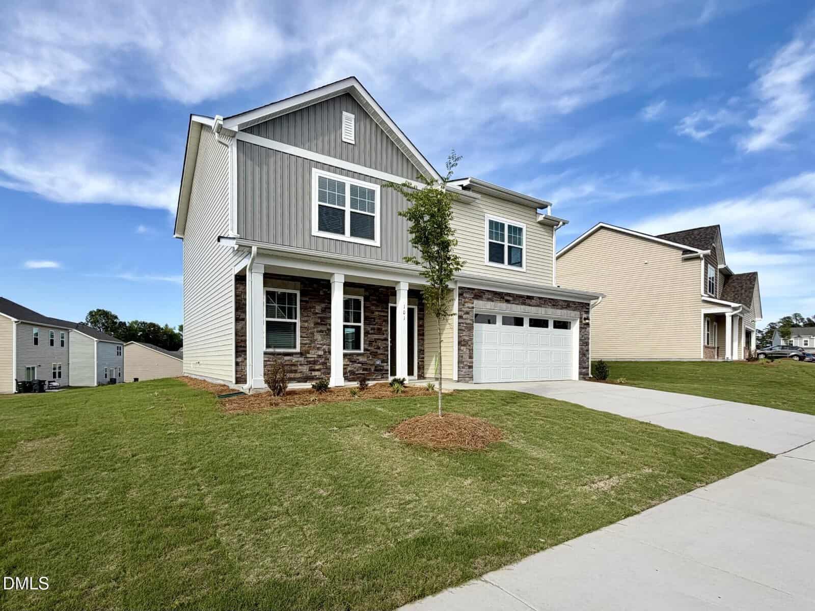 A modern two-story house with gray siding, stone accents, a covered front porch, and a two-car garage, surrounded by a neatly manicured lawn and a young tree, under a blue sky with scattered clouds.