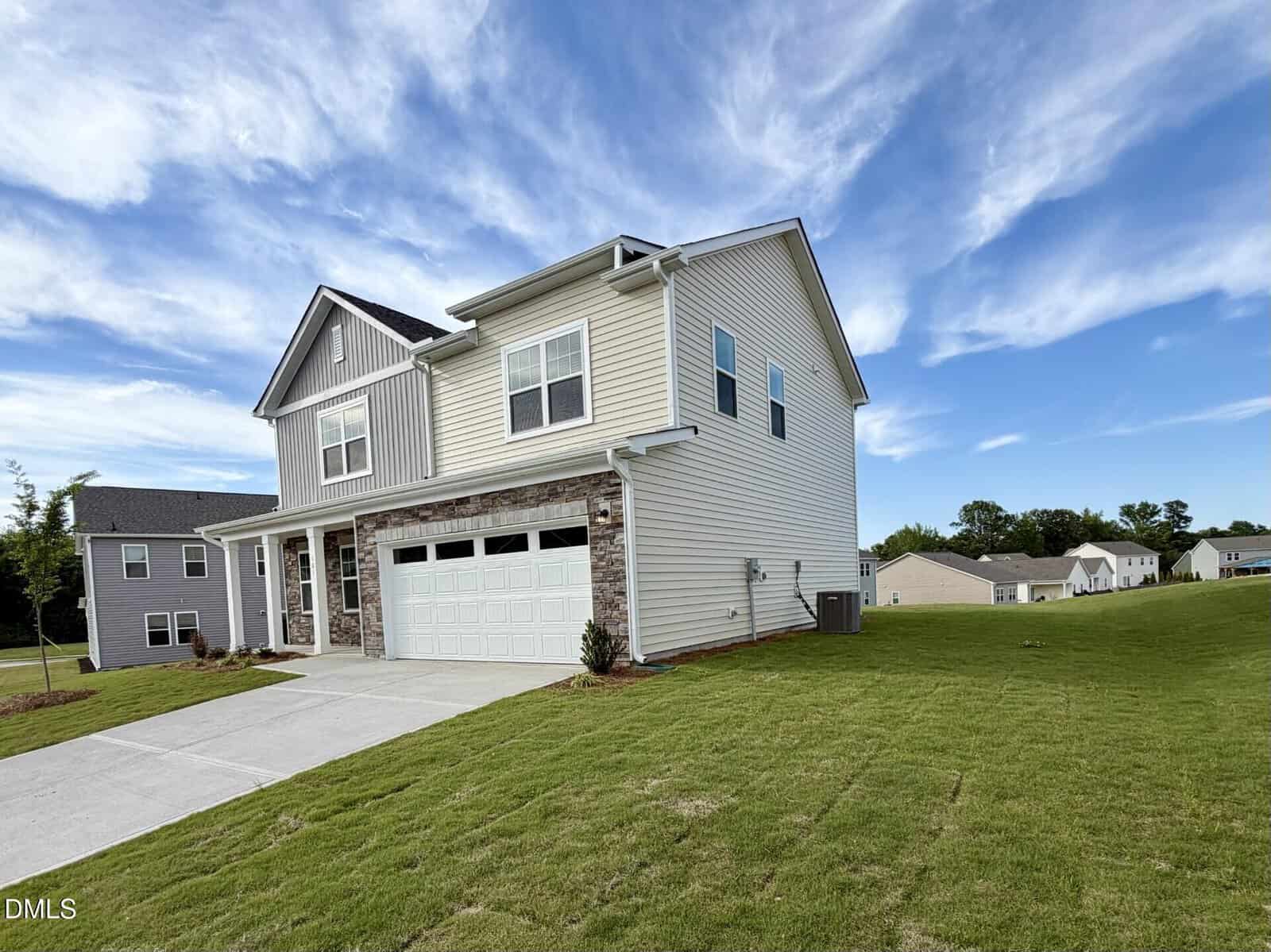 A two-story suburban house with beige siding, stone accents on the garage, and large windows, situated on a neatly trimmed lawn under a blue sky with scattered clouds.