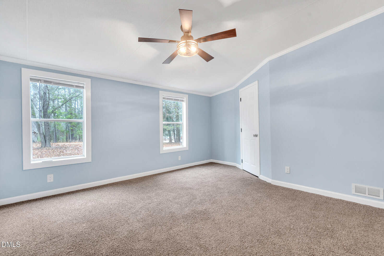 Empty room with light blue walls, two windows, brown carpet, a white door, and a ceiling fan with a light fixture; trees are visible outside the windows.