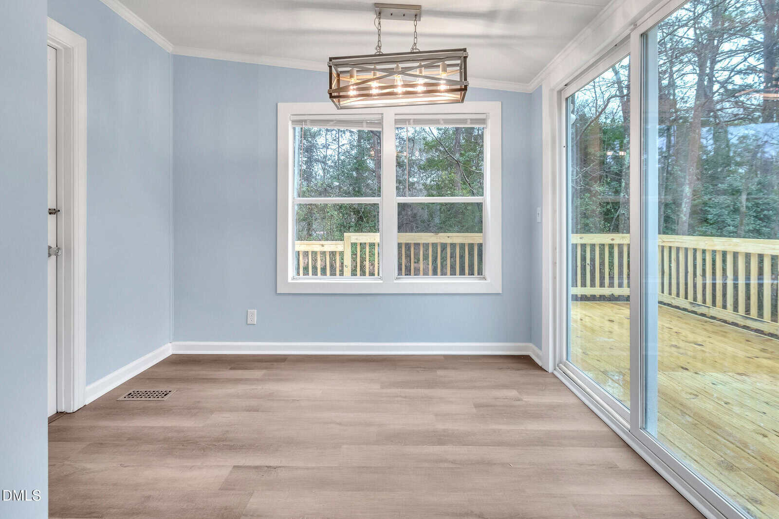 A small, empty room with light blue walls, wood-look flooring, a modern rectangular ceiling light, a window with a view of trees, and a large sliding glass door leading to a wooden deck outside.