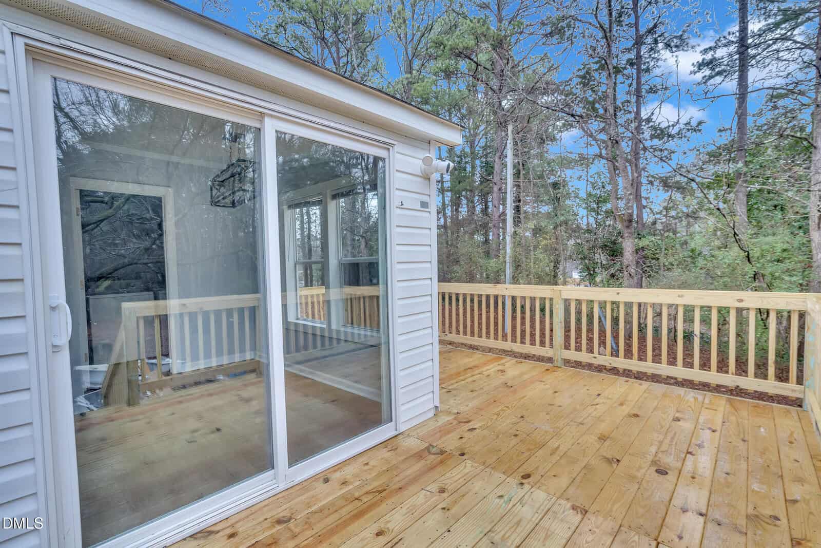 A wooden deck with a railing is attached to a white house. Large sliding glass doors lead from the deck into the house. Trees and a clear blue sky are visible in the background.