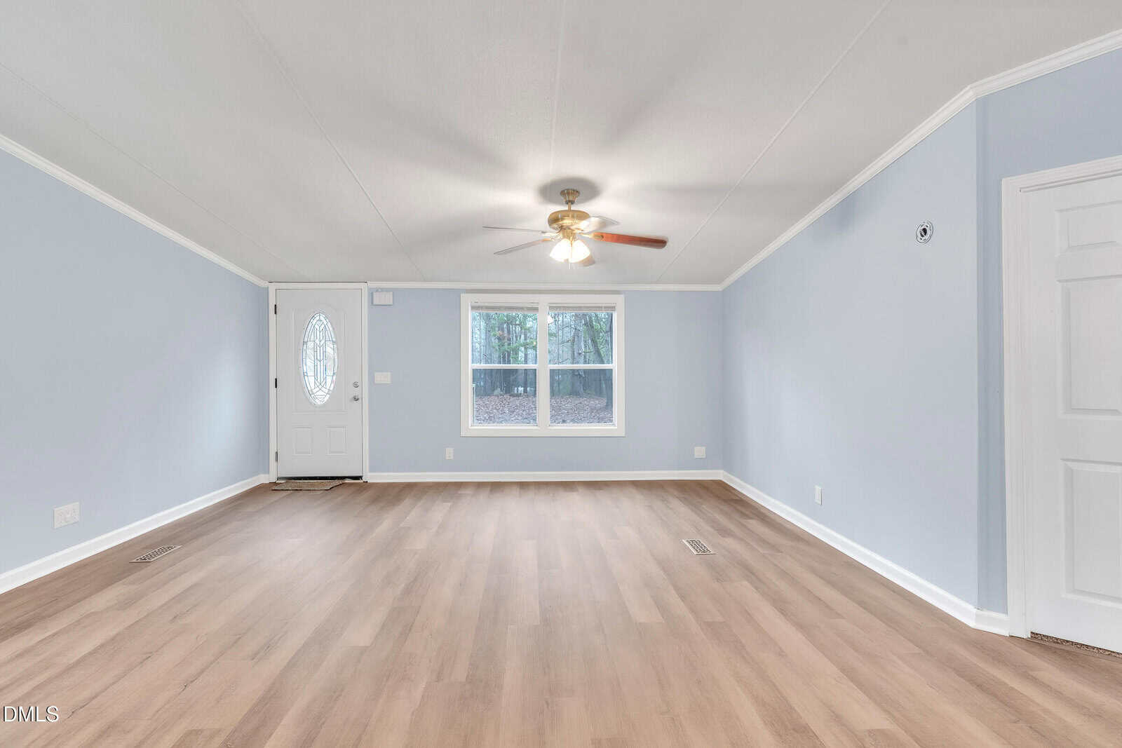 A bright, empty living room with light wood flooring, pale blue walls, a ceiling fan with lights, a white front door with an oval window, and two windows letting in natural light.