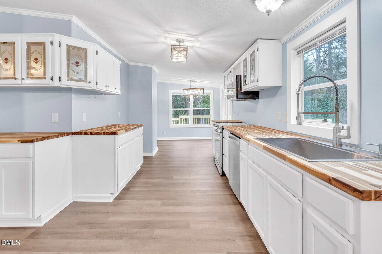A spacious kitchen with white cabinets, butcher block countertops, a large sink, stainless steel dishwasher, light blue walls, and wood-look flooring. Windows provide natural light and offer a view of greenery outside.