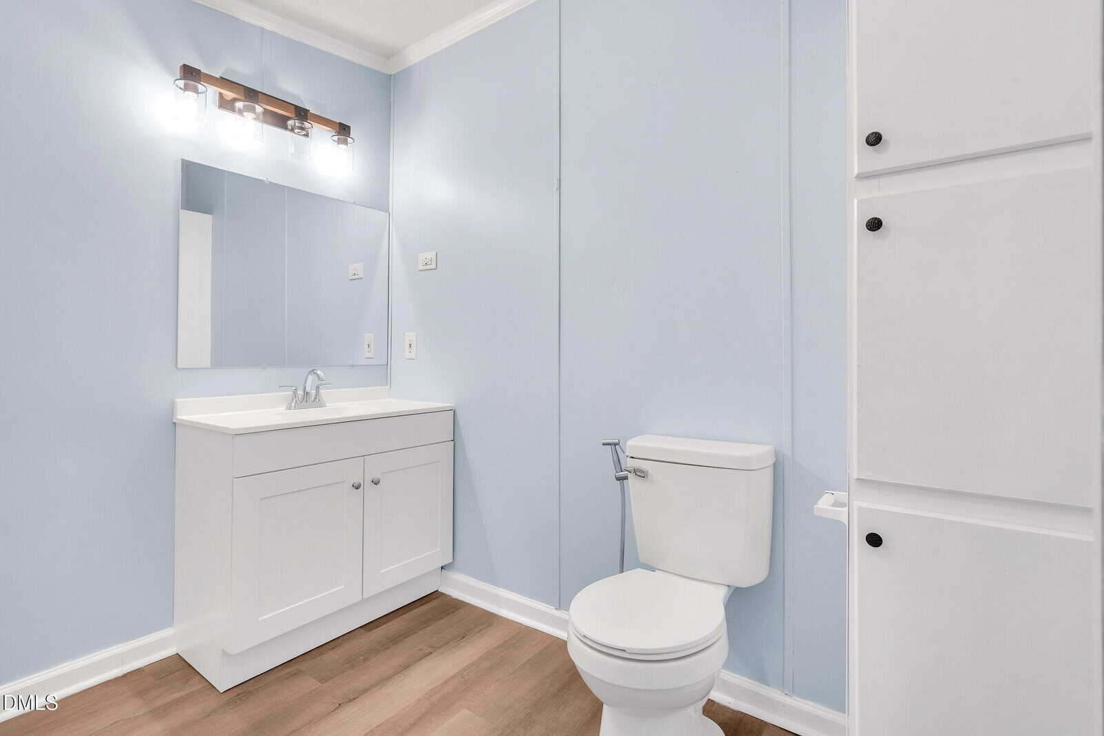 Bathroom with light blue walls, a white vanity with a mirror and light fixture above it on the left, a white toilet in the center, and built-in white storage cabinets on the right. Floor is light wood.