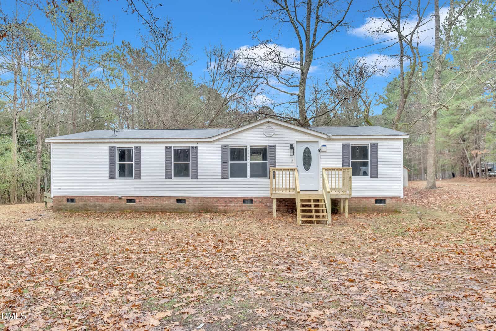 Located at 366 Buckskin Road in Henderson, this white single-story manufactured house features a brick foundation, multiple windows, and a small wooden front porch. The yard is blanketed with fallen leaves and surrounded by trees under a blue sky.