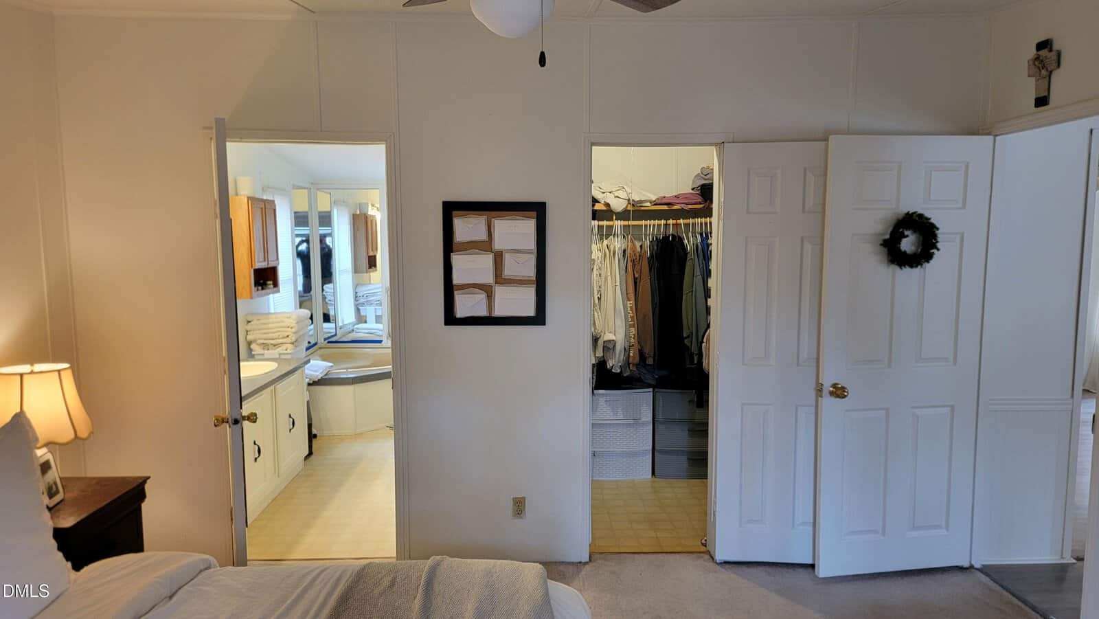 Bedroom view shows two open doors; one leads to a bathroom with a bathtub, vanity, and cabinets, and the other to a walk-in closet with clothes and storage bins. A closed door with a wreath is on the right.
