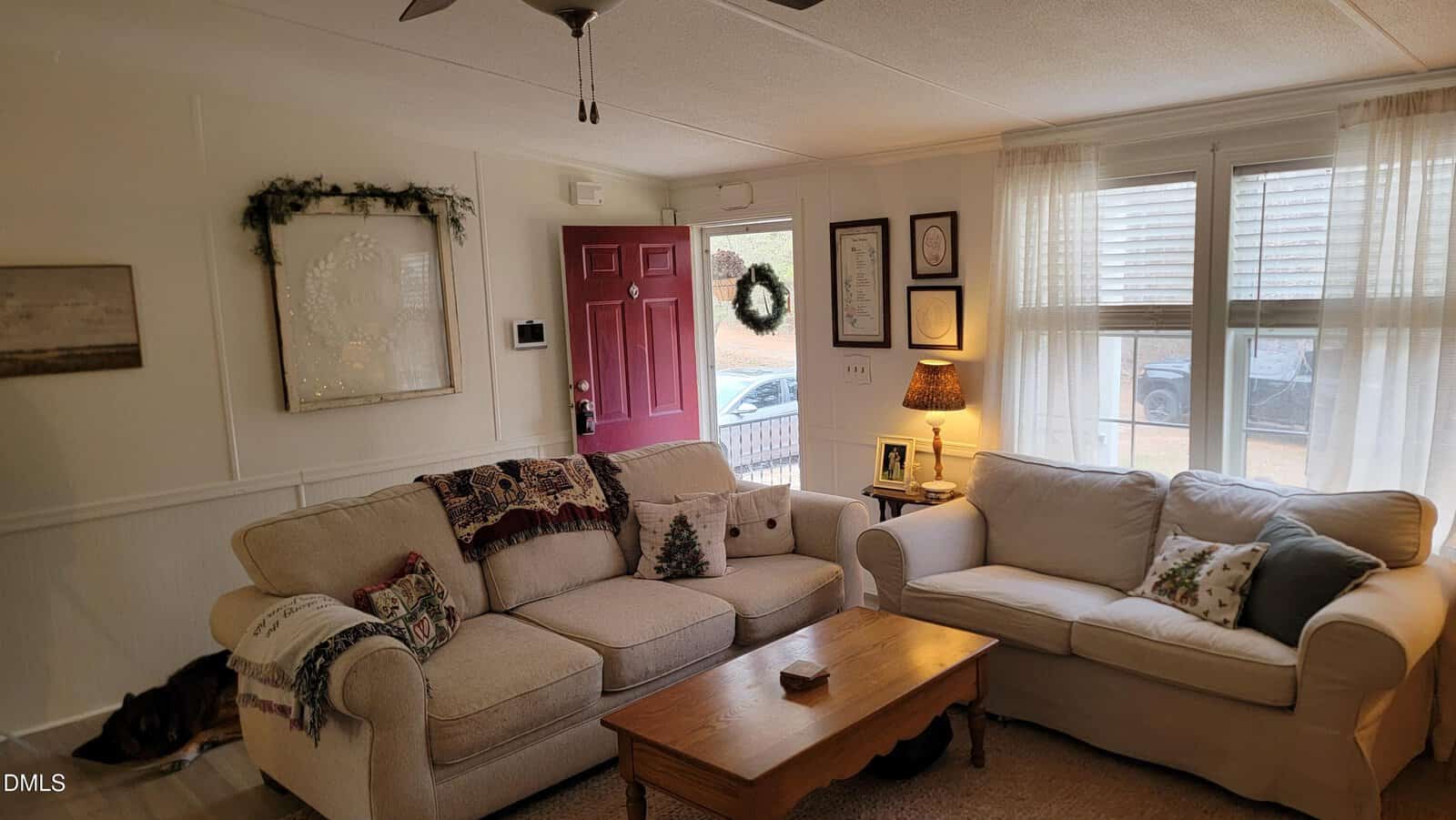 A living room with two beige sofas, a wooden coffee table, a red front door, framed art and wreaths on the walls, a small lamp on a side table, and light coming through white curtains on the windows.
