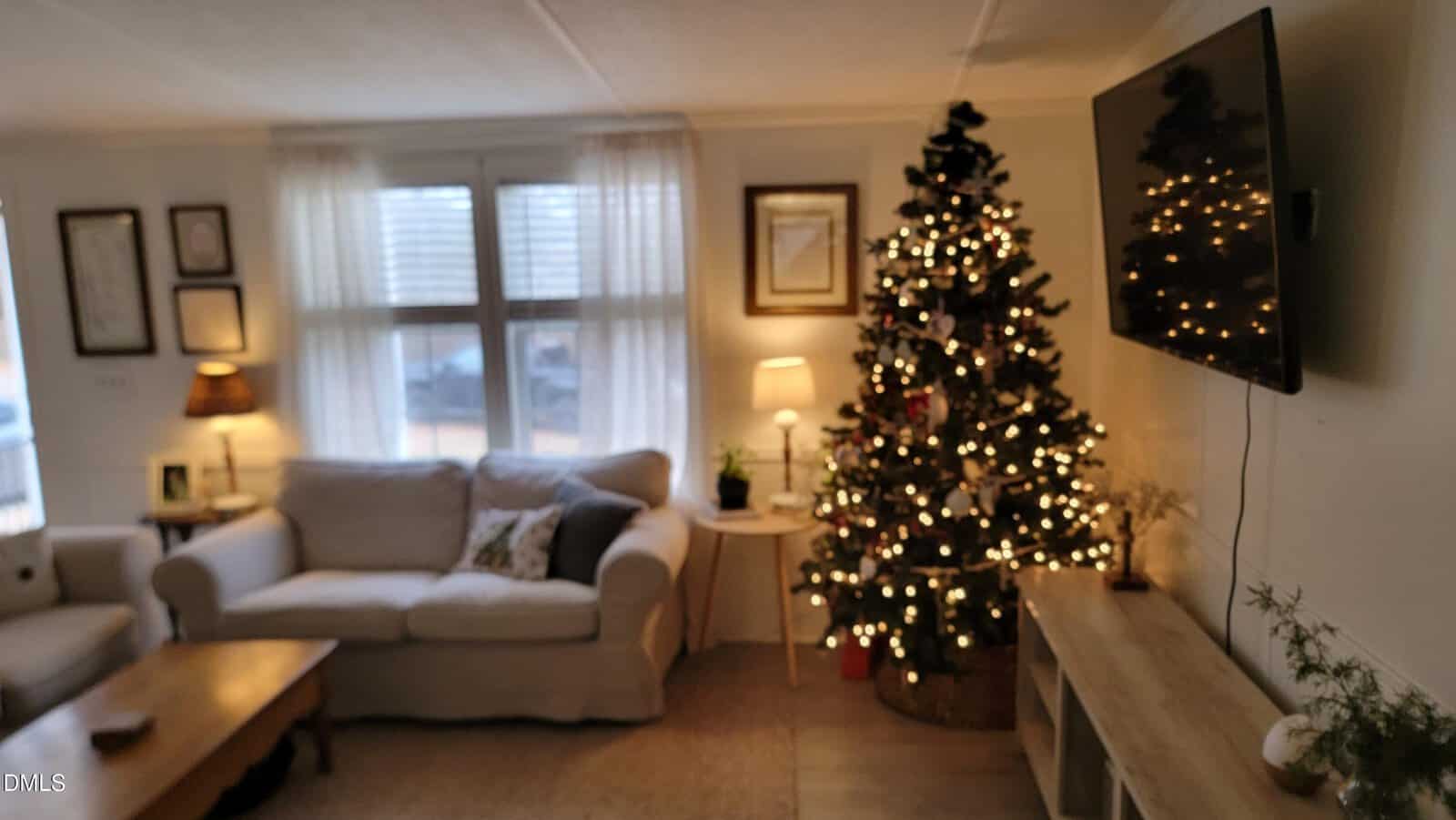 A living room with a lit Christmas tree next to a white sofa and a window with sheer curtains. A large flat-screen TV is mounted on the wall, and warm lighting illuminates the space.