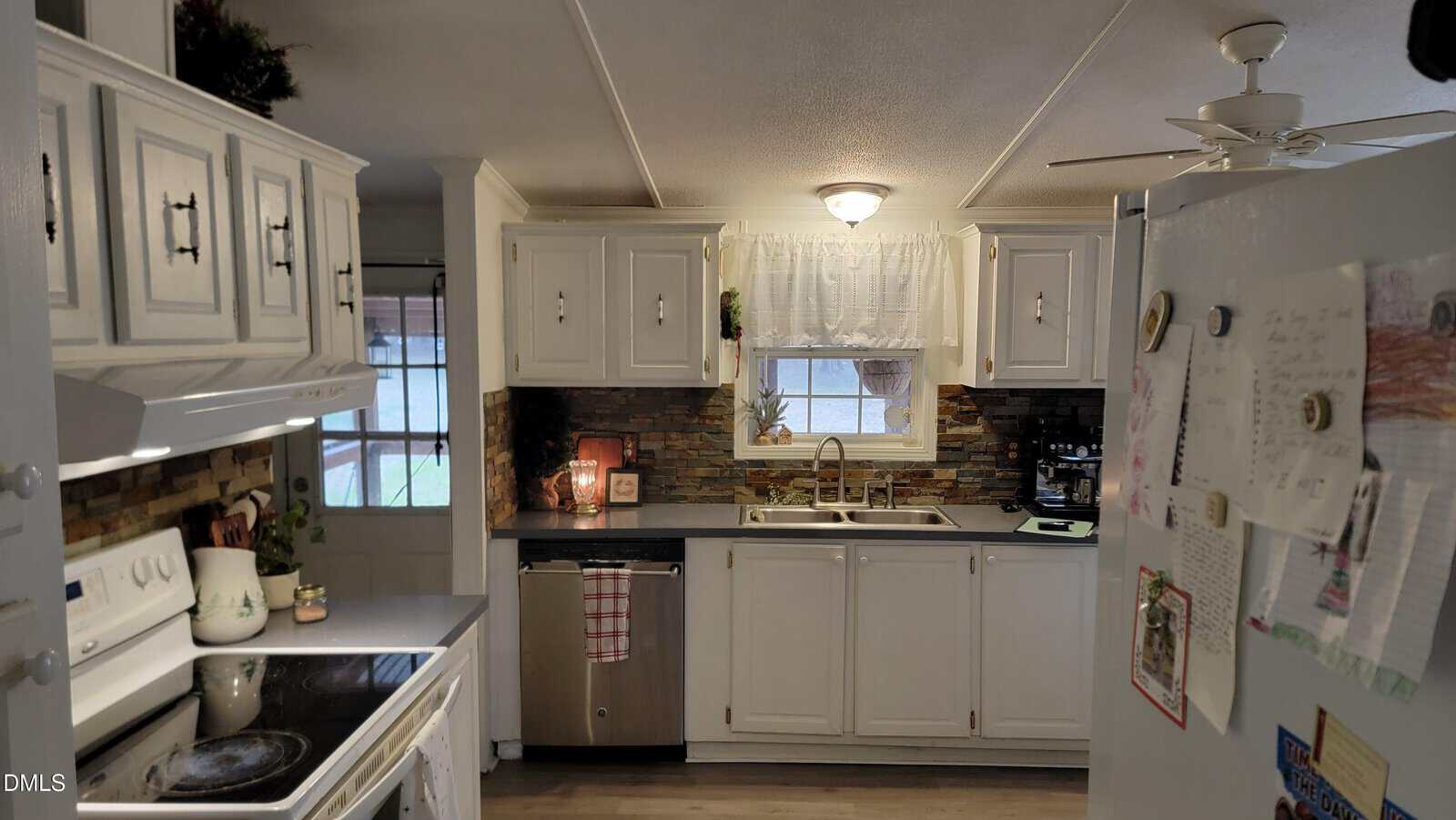 A kitchen with white cabinets, a white stove, a sink under a window with a curtain, a tiled backsplash, a refrigerator with papers and magnets, and various small appliances and decor on the counters.