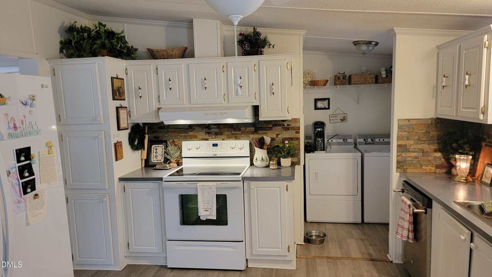 A white kitchen with an electric stove, white cabinets, and stone backsplash. The kitchen opens to a laundry area with a washer, dryer, shelves, and small appliances. There are plants and decor on cabinets.