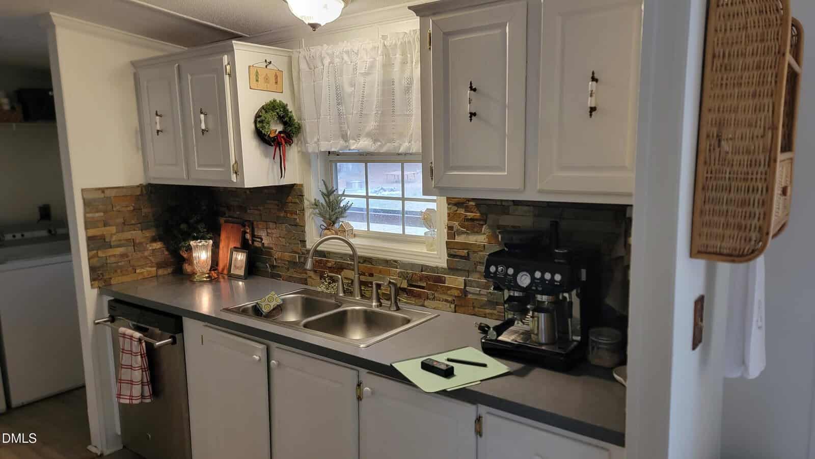 A cozy kitchen with white cabinets, stone backsplash, a double sink, coffee maker, stove, and kitchen decor. A window with sheer curtains lets in light above the sink, and a green cutting board sits on the counter.