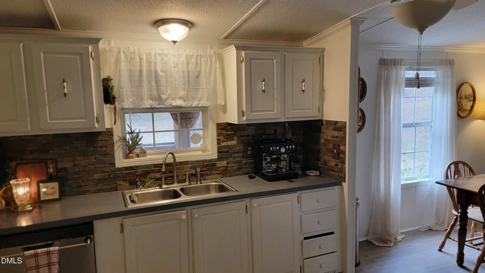 A cozy kitchen with white cabinets, a double sink under a curtained window, a coffee maker on the counter, stone backsplash, and part of a dining area with a wooden table and chairs by a window.