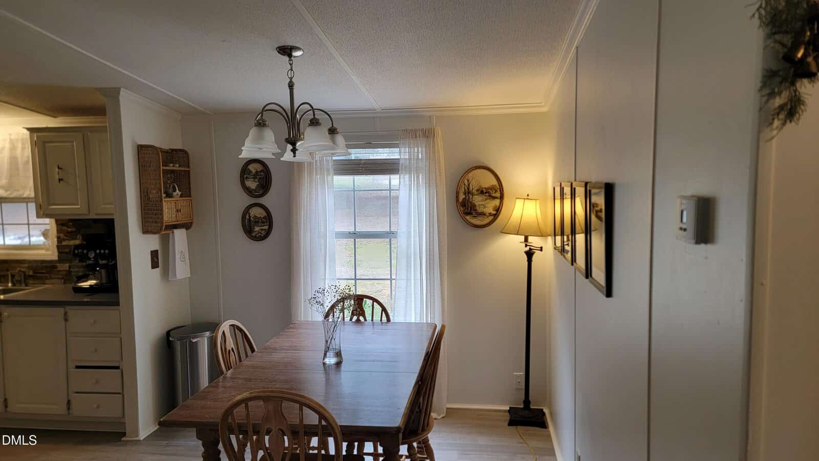 A dining area with a wooden table and four chairs, a hanging light fixture, wall art, a standing lamp, and a window with white curtains. The kitchen is partially visible to the left.