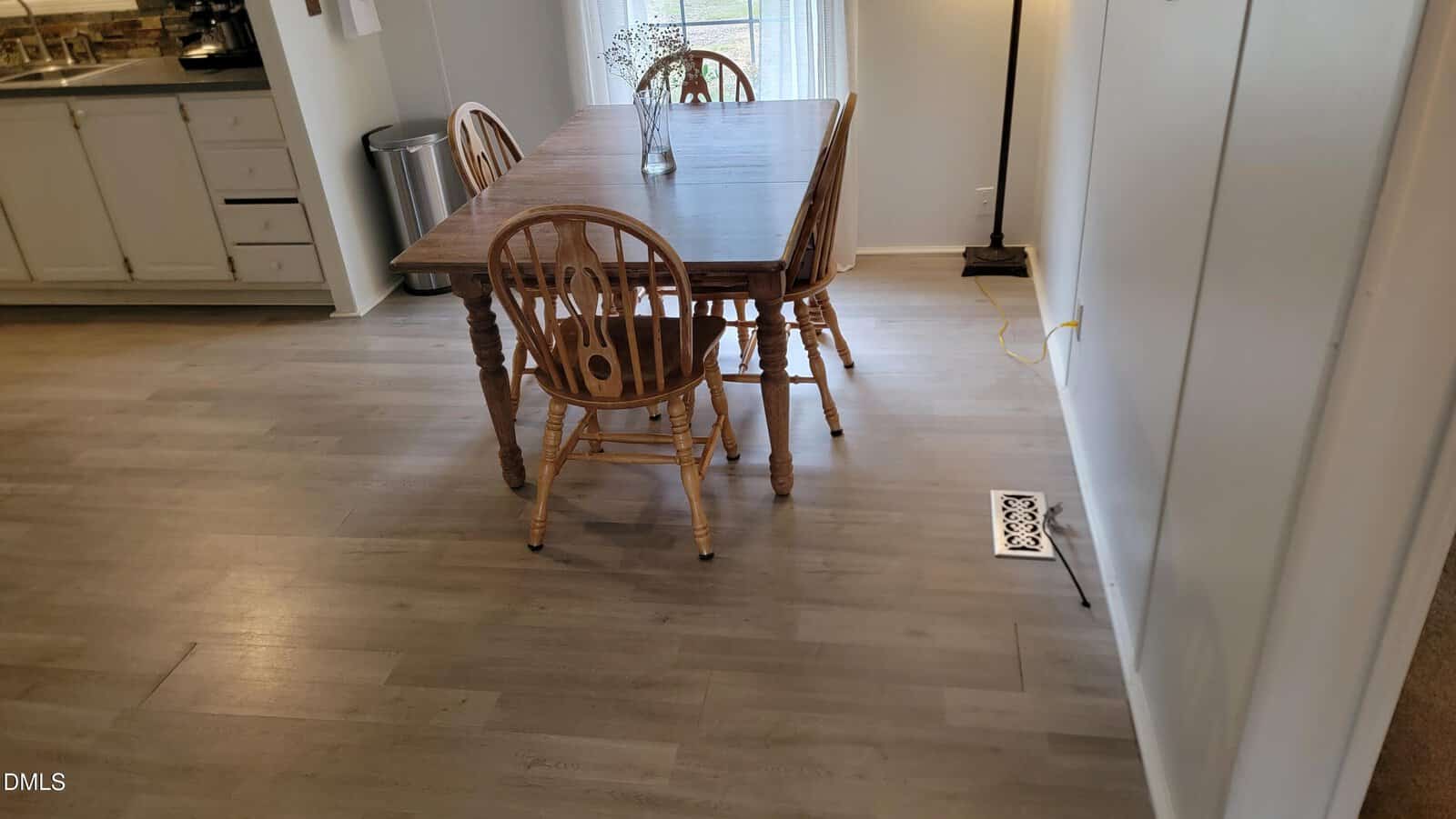 A wooden dining table with four matching chairs is placed on light wood flooring in a dining area next to a white wall. The kitchen with white cabinets is visible to the left. A window with sheer curtains is in the background.
