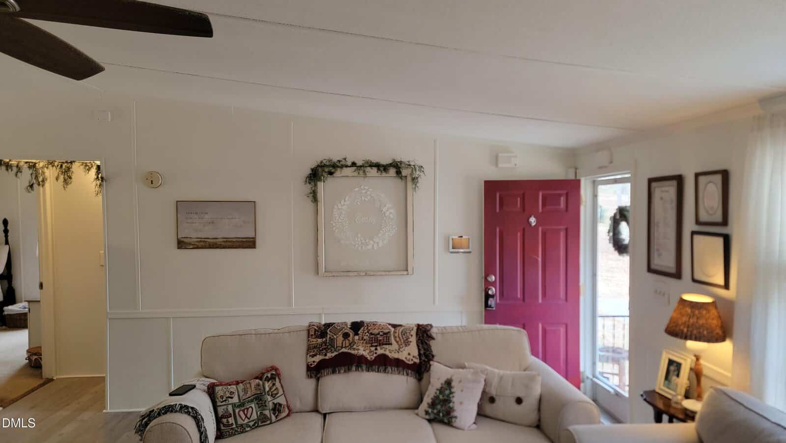 A living room with a cream-colored couch, decorative pillows, and a patterned blanket. A red front door is open, and framed art, a wall wreath, and a lamp are visible. Light enters through a window on the right.