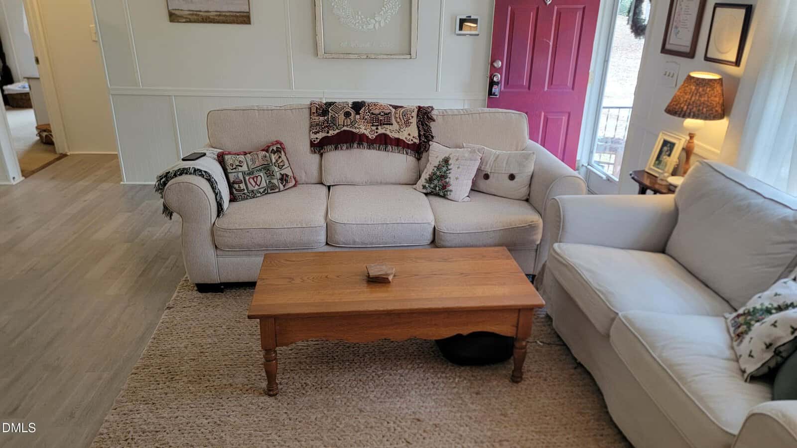 A living room with a beige sofa and a white couch, a wooden coffee table, a woven rug, and a red front door. The sofa has patterned throw pillows and blankets. A lamp sits on a side table near the window.