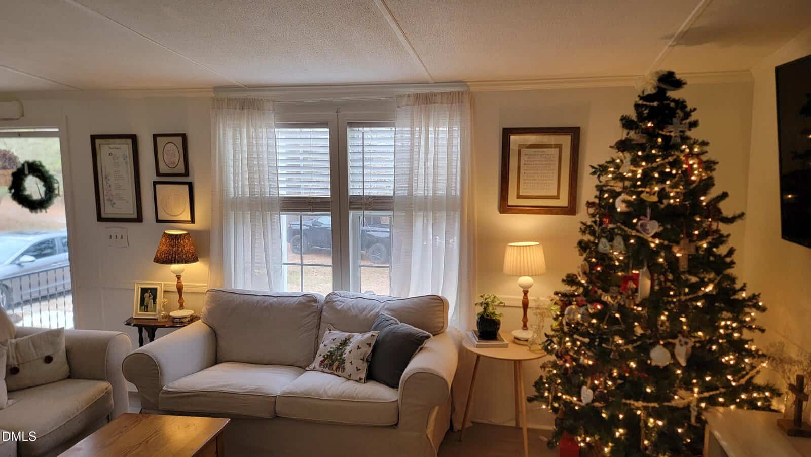A living room with a white sofa, side tables with lamps, framed art on the walls, a decorated Christmas tree, and a window with sheer curtains. A car is visible outside the window.