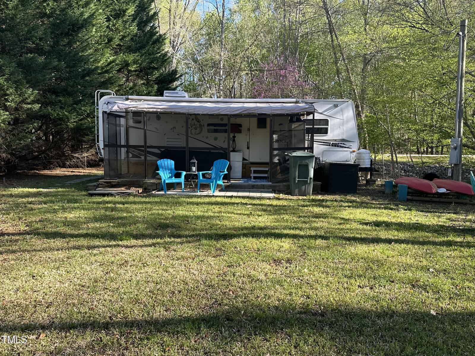 A trailer with a screened porch stands on a grassy lawn at 1507 Sagamore Drive in Louisburg, flanked by two blue chairs and a table. Trees and canoes are in the background, with sunlight filtering through the foliage.