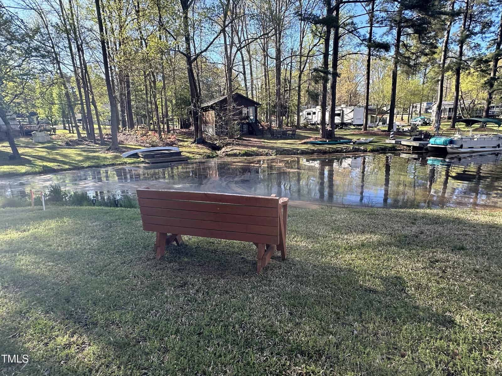 A wooden bench faces a small pond surrounded by grass and trees. In the background, there are boats, a dock, a small wooden shed, and several RVs or campers among the trees.