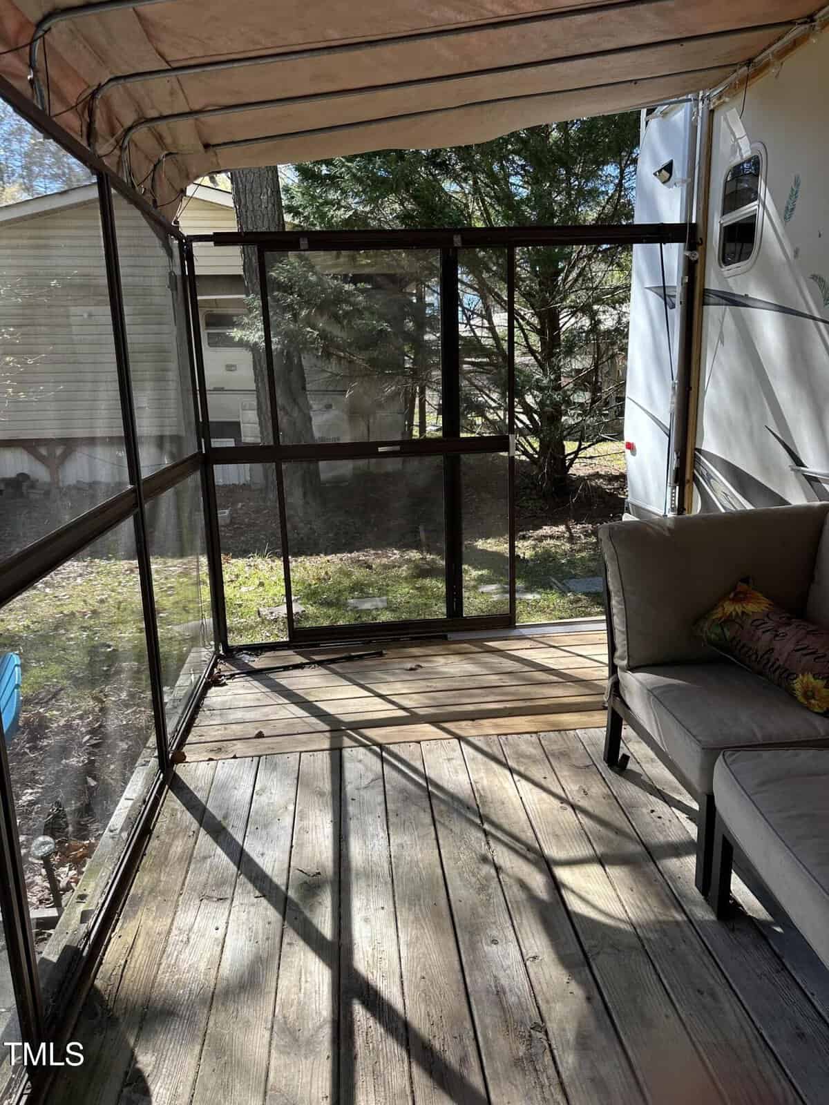 Screened patio area with wooden floor, partial view of a cushioned outdoor sofa, and sunlight casting shadows. Trees and an adjacent building are visible through the screen panels. A canopy covers the area.