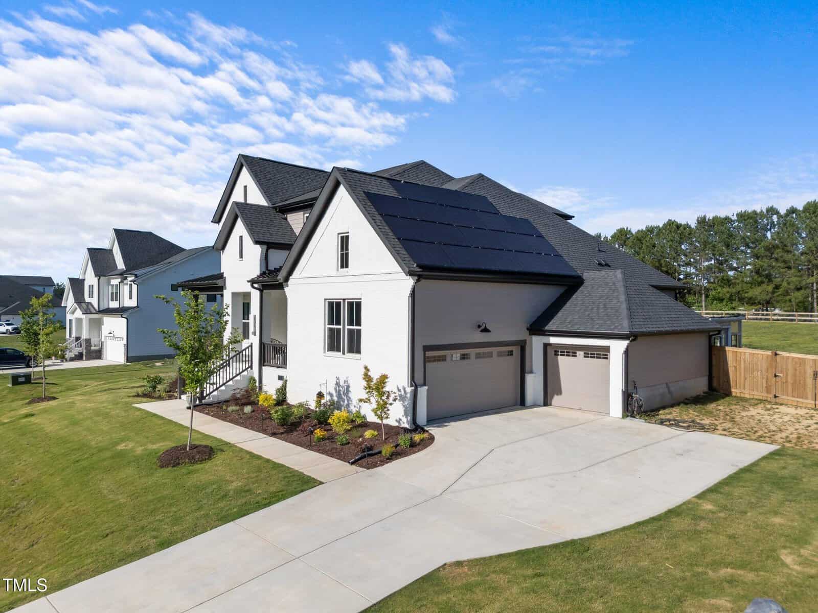 A modern two-story white house with black trim and solar panels on the roof, featuring a double garage, landscaped yard, and driveway under a blue sky with scattered clouds.